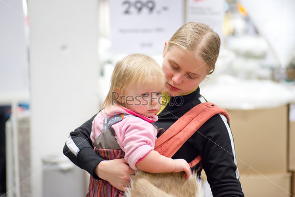 Young mother with daughter in sling shopping skin carpet in supermarket