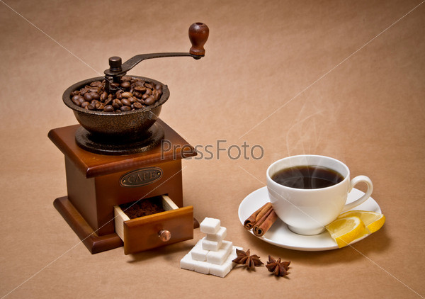 Coffee-grinder, cup of hot coffee, coffee beans, sugar, cinnamon sticks, two lemon slices and some star anise fruits  on extra-strong paper background