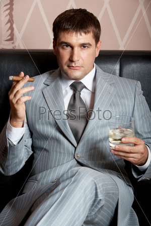 Portrait of handsome man in grey suit sitting on leather sofa with whisky and cigar in casino