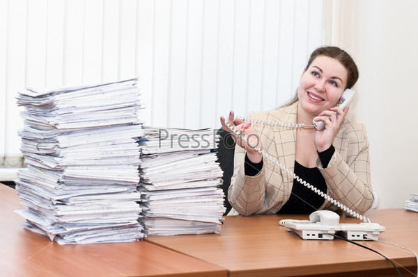 Young caucasian woman in office interior calling by telephone and stack of paper documents