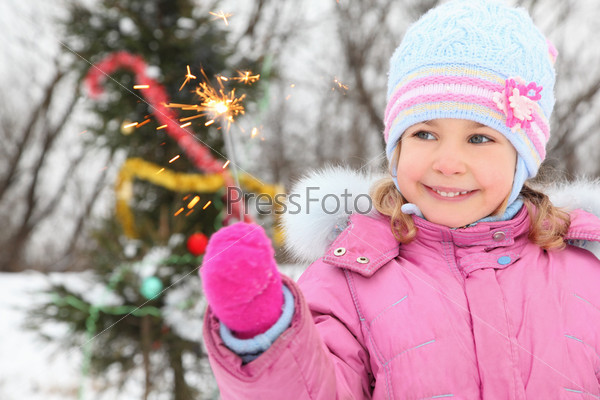 little girl wearing winter jacket is standing near christmas tree with bengal light. christmas tree in out of focus.