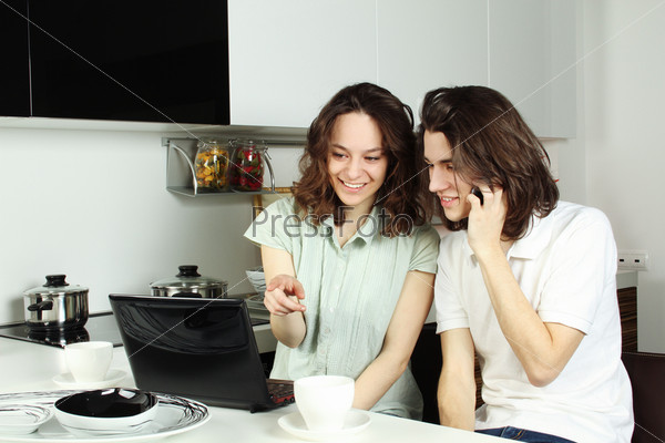 Young couple in the kitchen drinking coffee and working on a laptop, both good mood. The guy on the phone