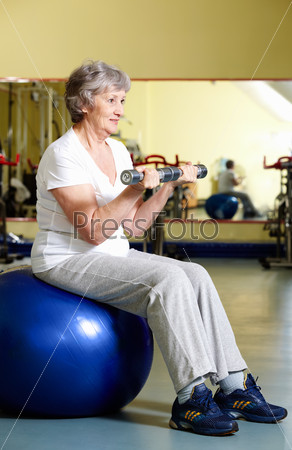 Portrait of aged woman doing physical exercise with barbells while sitting on blue ball