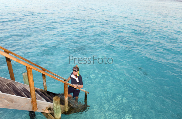 Man stands with equipment for Snorcel on steps of villa on piles on water.  Maldives.