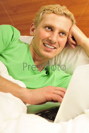 Attractive man relaxing in bed. White silk linen. Working by is laptop at home.