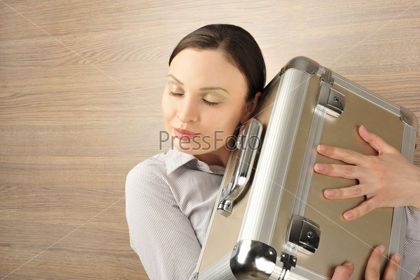 Portrait of emotional pretty young woman against modern stylish wooden wall holding big metal case. Embracing and dreaming