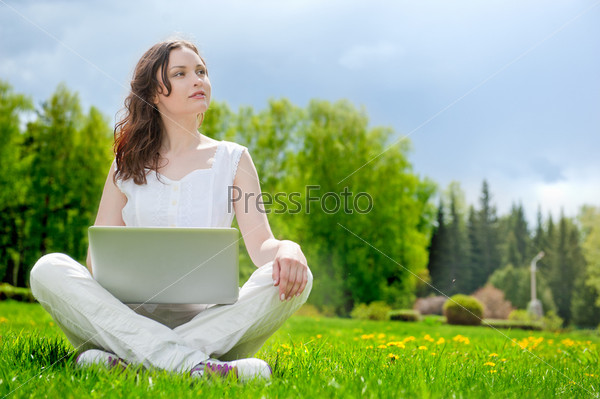 Young woman with laptop sitting on green grass and looking to a sky