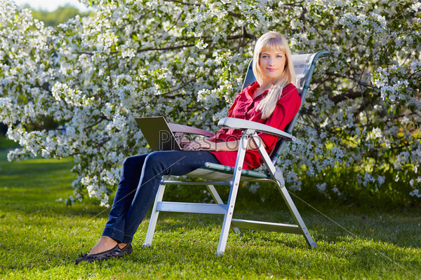 outdoor portrait of beautiful blonde girl sitting in folding chair on green grass near bloomin apple tree with laptop