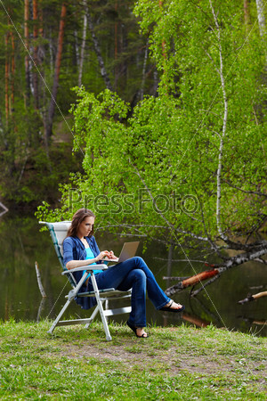 outdoor portrait of beautiful woman sitting in folding chair with laptop near the pond in forest