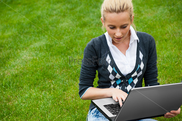 Young woman with laptop sitting on green grass and looking to the screen of laptop