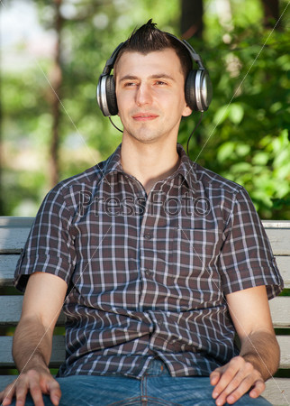 Portrait of a relaxed young man sitting on bench in park and listening to music on headphone