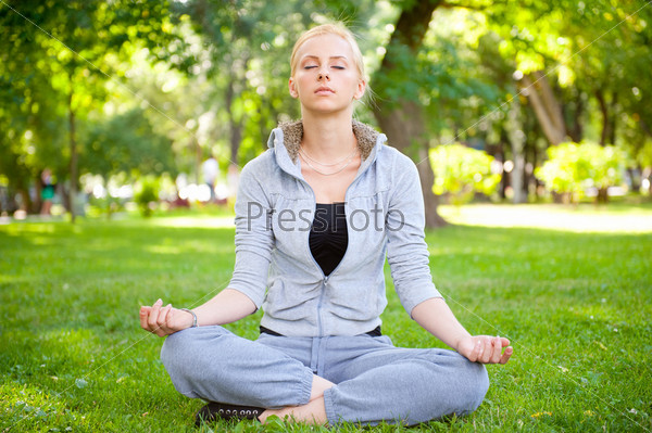 Portrait of young woman meditating in pose of lotus on green grass on meadow at summer park under tree