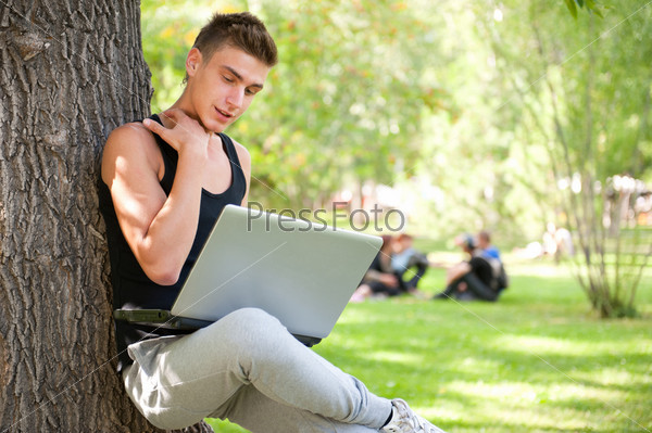 Closeup portrait of young student man using laptop leaning on tree trunk on foreground at summer park. Group of students sitting on the grass at the background