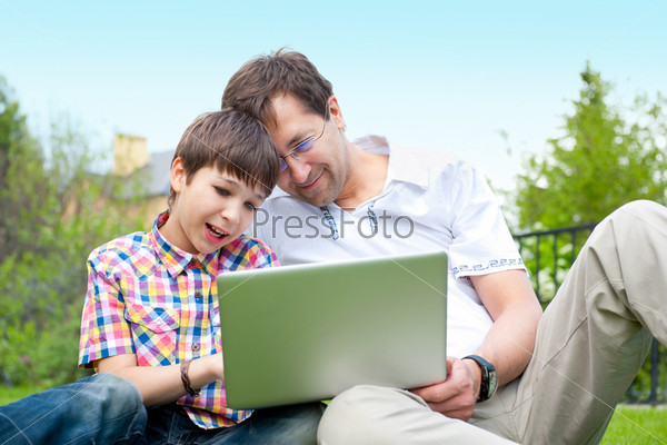 Closeup portrait of happy family: father and his son using laptop outdoor at their backyard sitting on the grass together
