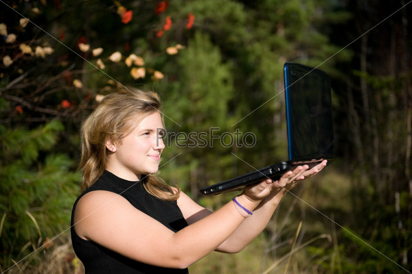 The girl holds the laptop on hands. There is it on the nature. The girl smiles.