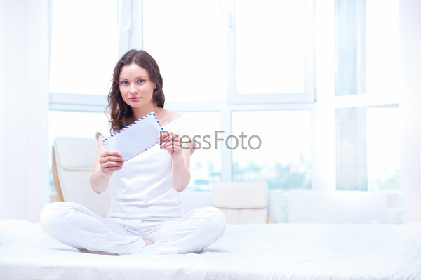 American or european young pretty woman sitting on bed in bright bedroom and opening letter. She is excited and dreaming about enrollment to her favorite college or university.