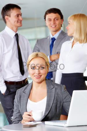 Pretty businesswoman looking at camera at workplace with several partners on background