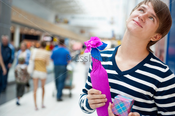 Portrait of young man inside shopping mall standing relaxed and holding gift box and bottle. He is preparing to visit his girlfriend and make a proposal