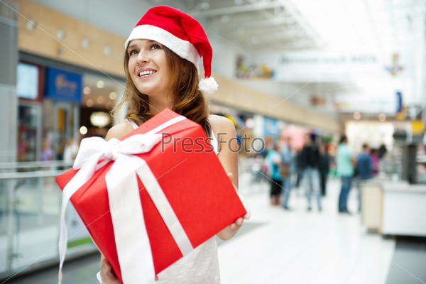 Excited attractive woman with big gift box standing at shopping mall wearing Santa Claus cap