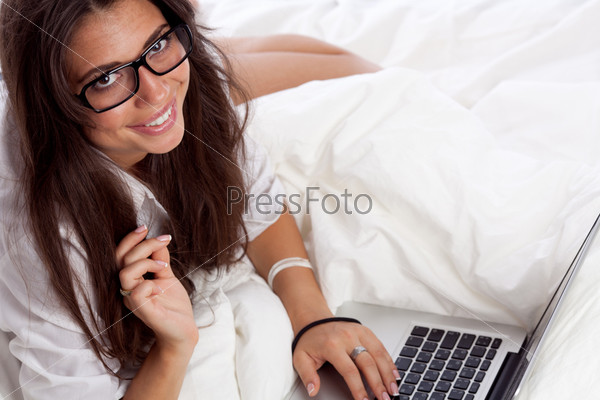 Молодая женщина в очках с ноутбуком лежит на кровати