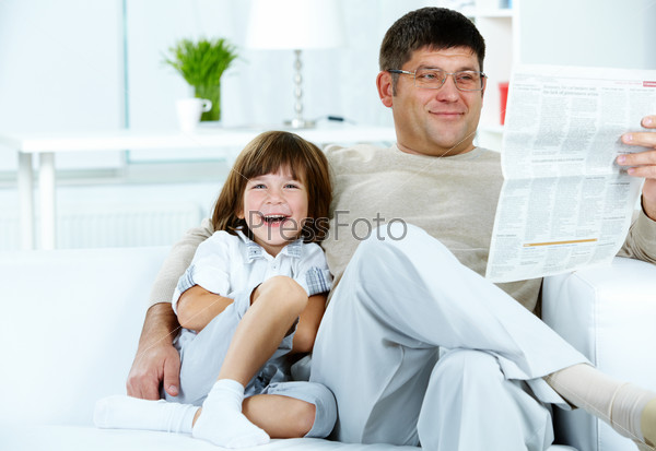 Photo of happy boy sitting by his father reading paper at home