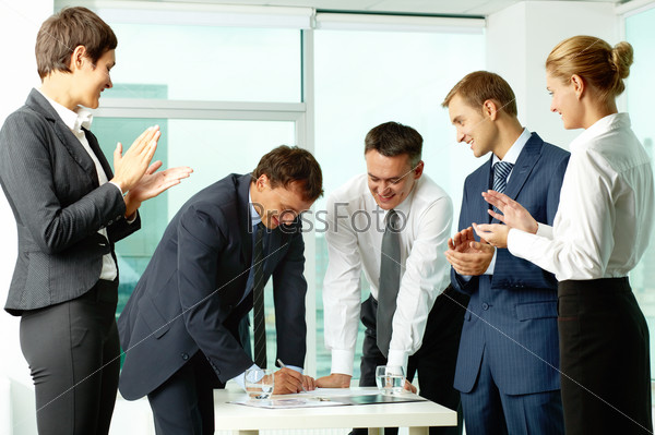 Image of co-workers congratulating their colleague while he signing paper at meeting