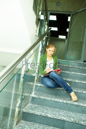 Photo of pretty girl sitting on stairs of college and writing something