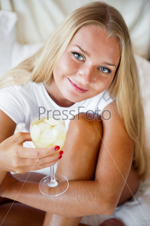 Woman drinking fresh ice tea beverage and looking at camera while sitting on her bed at home