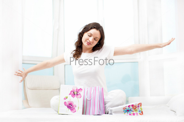 Portrait of young beautiful awake woman with gifts on bed at bedroom. She is happy and daydreaming