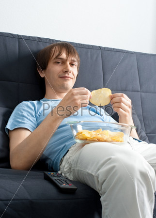 Молодой парень смотрит телевизор и ест чипсы, сидя дома на диване