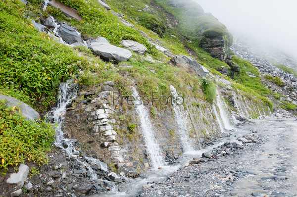 Waterfall on a mountain road with stone wall. Focus on the front of the wall.