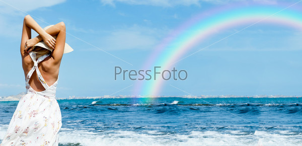 Young fashionable woman wearing hat and sophistical dress on the beach looking on rainbow. Photo from behind