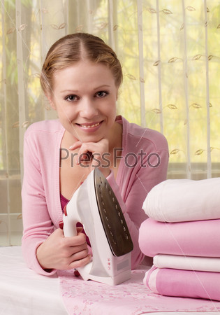 Счастливая молода женщина в розовом держит в руке утюг на гладильной доске с чистыми розовыми вещами