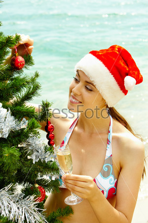 Portrait of female in bikini and Santa cap holding champagne flute and looking at decorated firtree on the beach