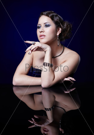 portrait of young beautiful brunette woman in bracelet, beads, ring and ear-rings at mirror table on blue