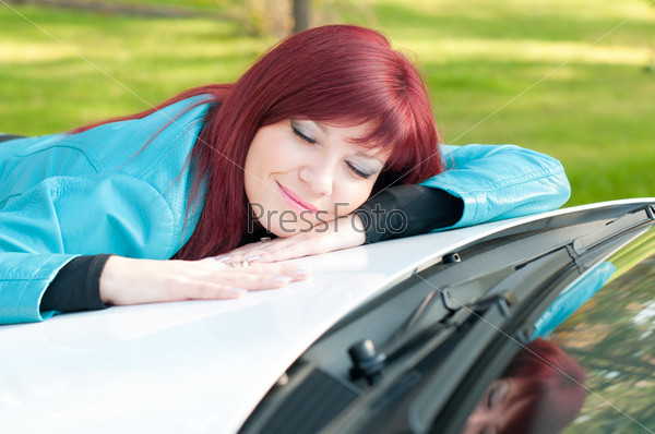 Happy red-haired young woman lying on the bonnet of her new car outdoors