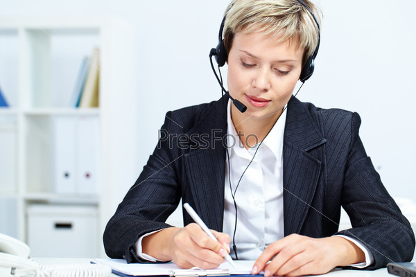 Portrait of young secretary with headset sitting at table and making notes