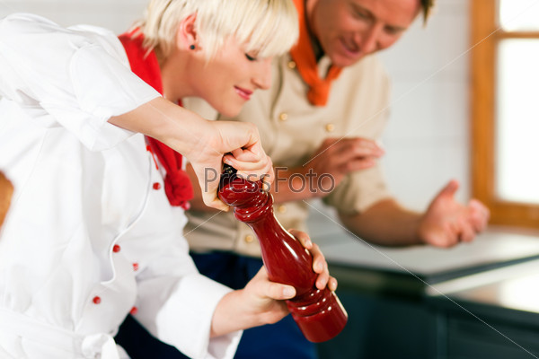 Two chefs in teamwork - man and woman - in a restaurant or hotel kitchen cooking delicious food, she is using a pepper mill (FOCUS ON MILL!)