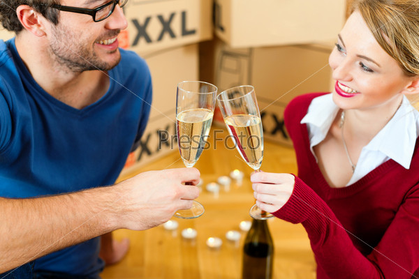 Young couple moving in a home or apartment, they are sitting on the wood floor drinking champagne having a break from renovation