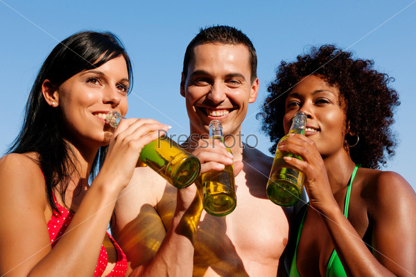 Group of friends - one man embraces two women and all have drinks in swimwear on the beach of a lake in summer