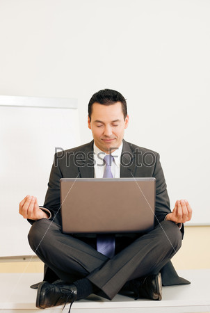 Office worker - a male manager - meditating cross legged upon his table, being very relaxed, a metaphor for work life balance