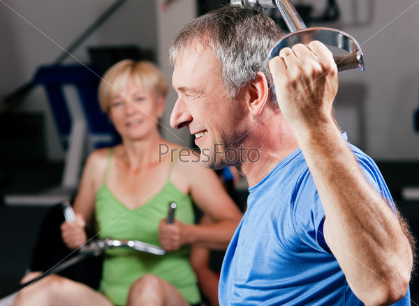 Senior couple -  man and woman - in the gym lifting weights on a lat pull machine, exercising
