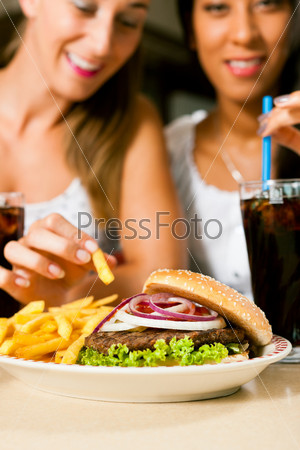 Two women - one is African American - eating hamburger and drinking soda in a fast food diner; focus on the meal, stock photo