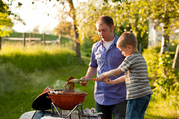 Family - father and son - having a barbecue party, the child is turning meat and sausages, both having fun