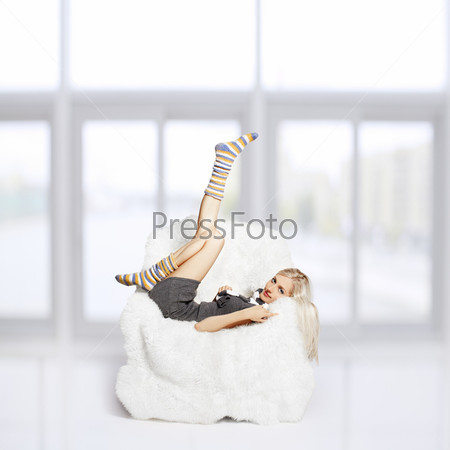 portrait of beautiful blonde sitting on big white furry arm-chair and putting on long striped socks