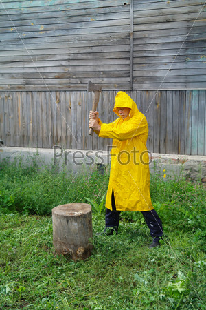 Пожилой мужчина в желтом плаще с топором на фоне деревенского дома