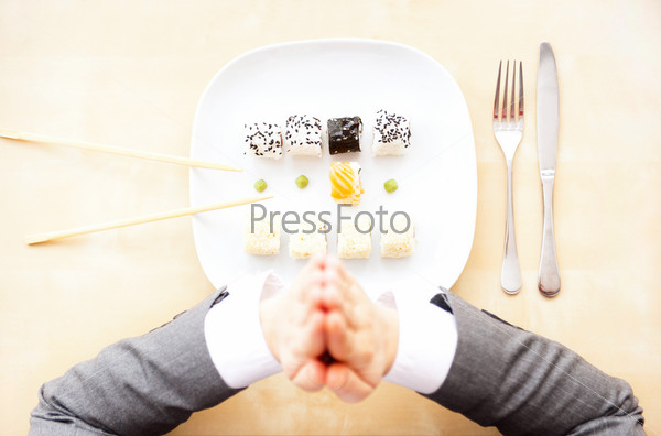 Top view of sushi set on white plate, sticks and fork and knife on other side, praying hands of businesswoman. European-Asian consensus and perfect sushi service concept