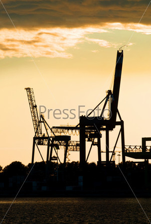 Silhouettes of two motionless, erect, idle harbor cranes against the setting sun