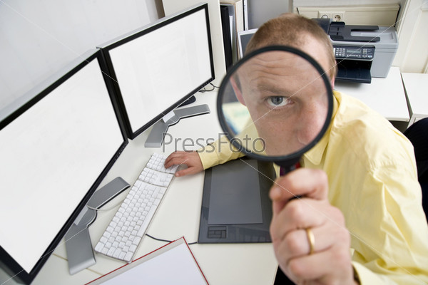 The eye of a businessman, sitting behind a dual screen computer, seen through a magnifying glass, illustrating being careful, watchful and shrewd in business