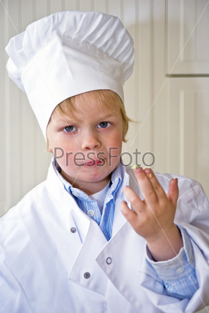 A boy, dressed in a chef\'s uniform, showing his dirty fingers, sticky with the sweet ingredients he\'s been tasting. Selective focus on the boys face